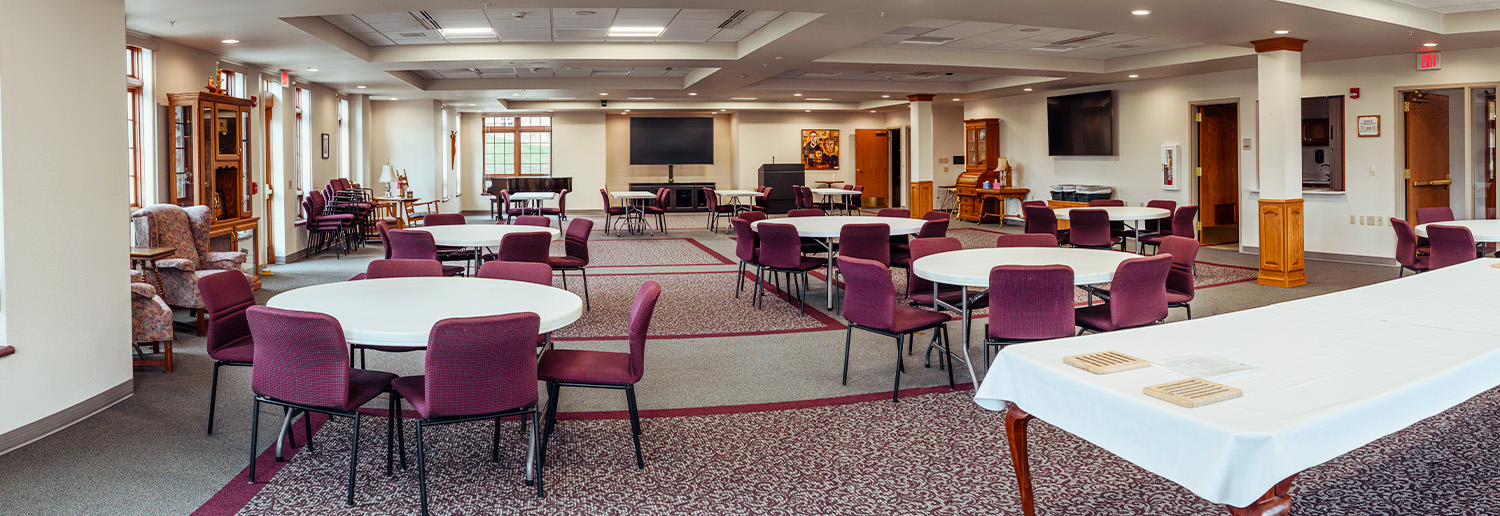 A photo of Founders Hall from the back of the room showing circular tables with maroon chairs and large screen at the front of the room.
